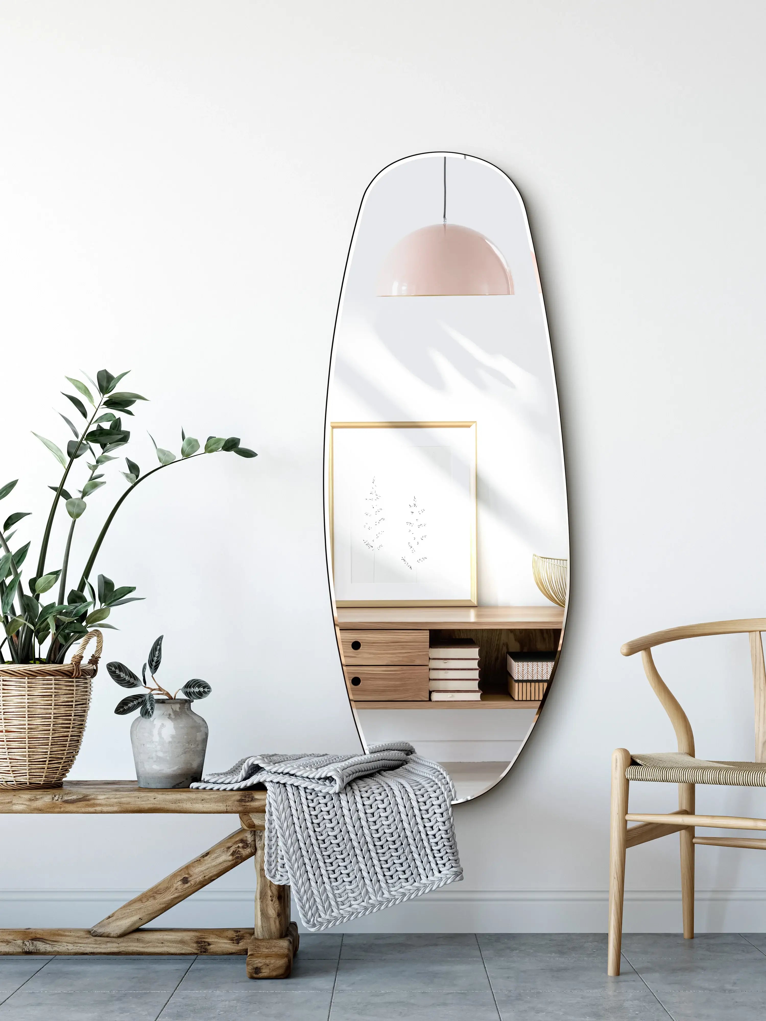 A modern entryway features a tall opal mirror, a wooden bench with a knit blanket, potted plants, a small wooden shelf with unique decor, and a pink pendant lamp reflected in the mirror.